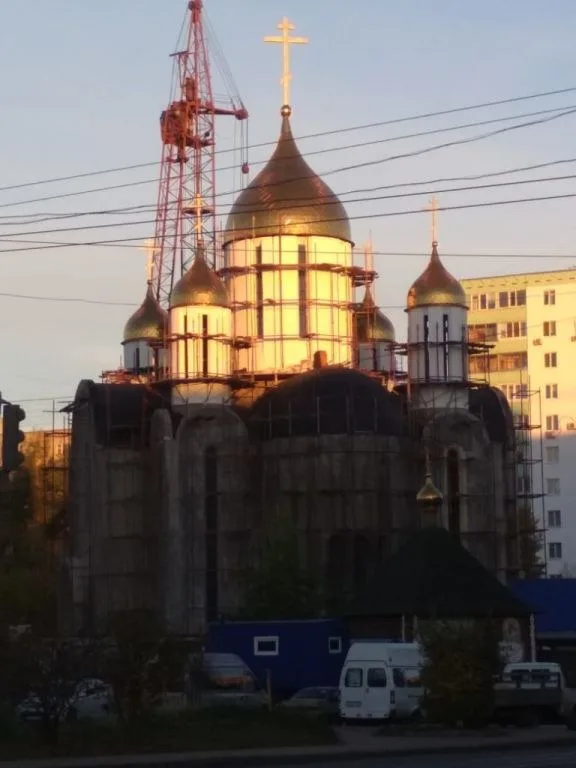 Недостроенный храм в строительных лесах, купола сияют золотом в закатном свете, рядом башенный кран