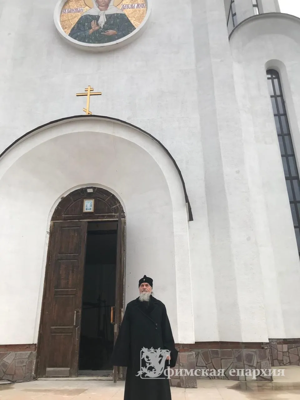 Священнослужитель у арочного входа храма с резной дверью и образом блаженной Матроны Московской над притвором