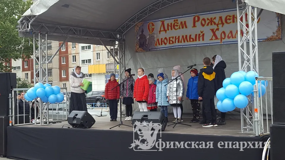 Дети в верхней одежде стоят у микрофонов на уличной сцене, украшенной синими шарами, за ними растяжка с поздравлением храму