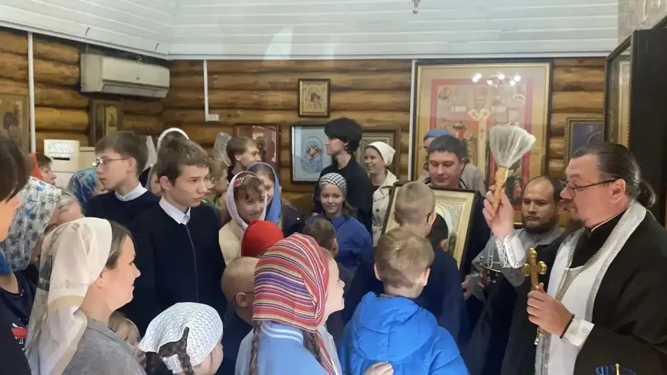 Группа детей и педагог слушают беседу в деревянном храме, на заднем плане иконы на стенах